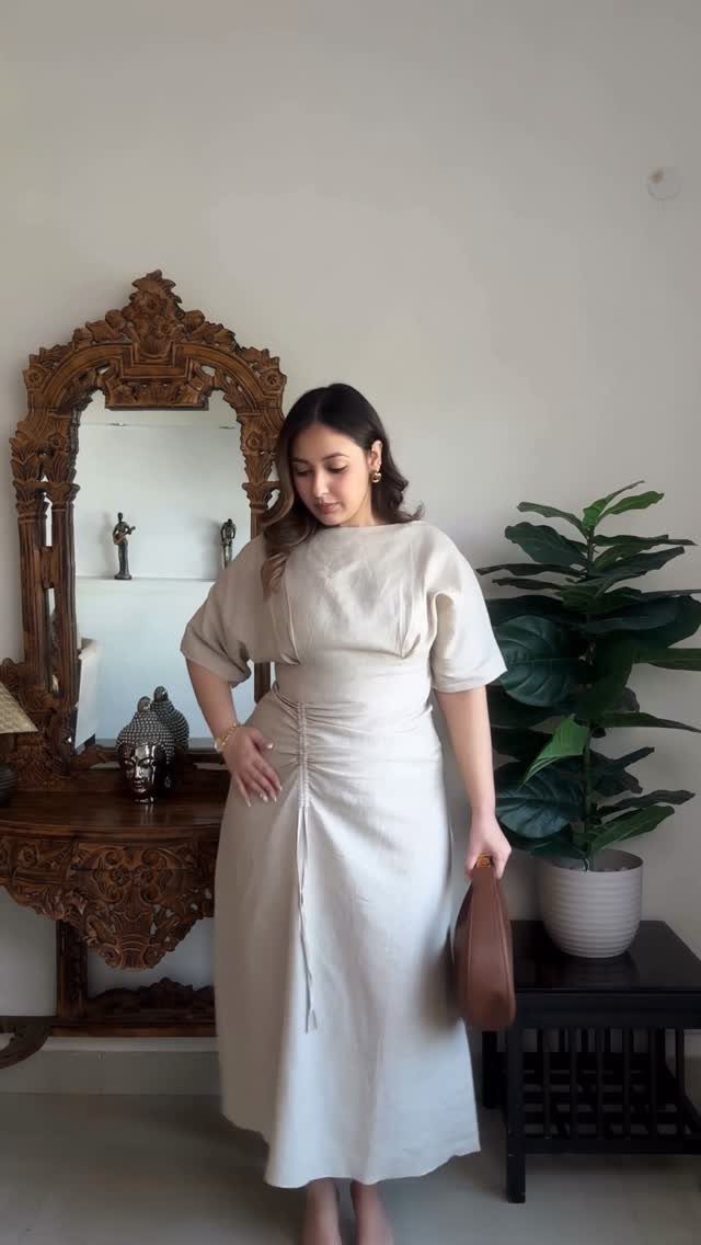 Woman in a cream midi dress standing in a room with an ornate mirror.