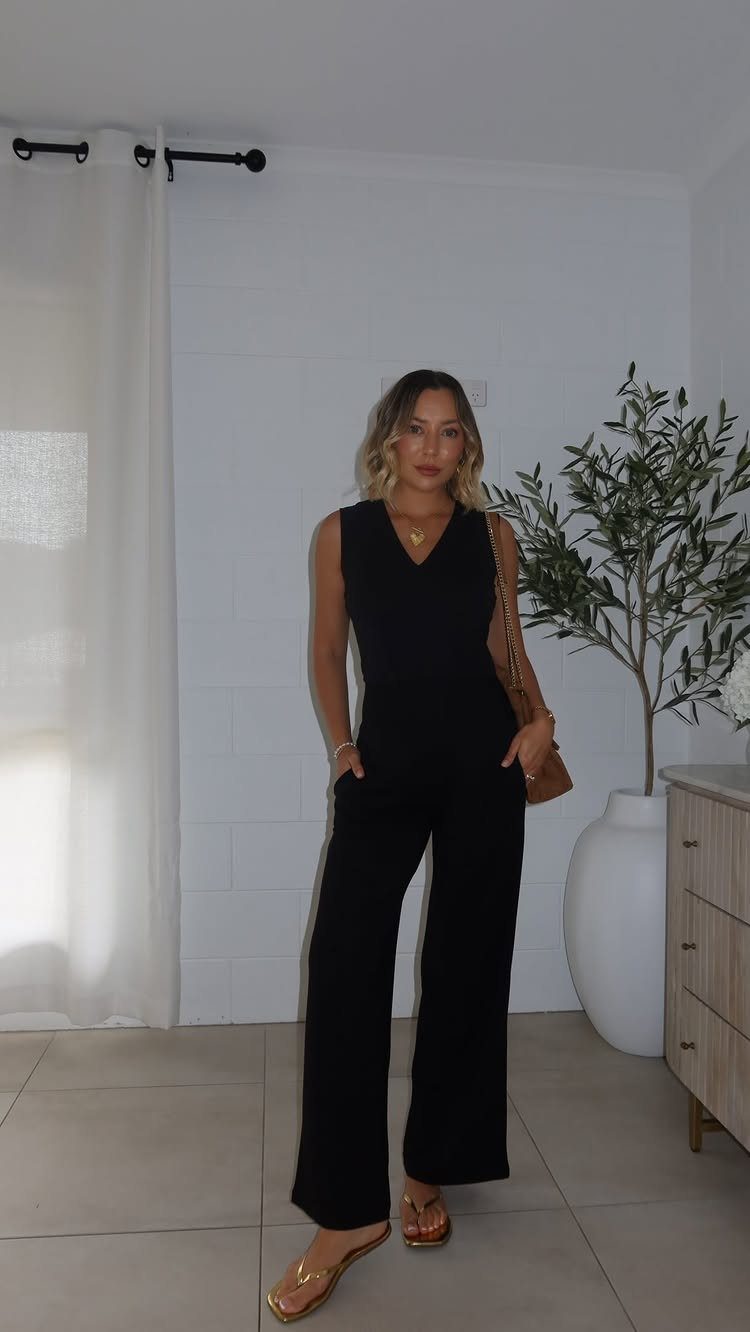 A woman posing in a black sleeveless wide-leg jumpsuit and gold jewelry.