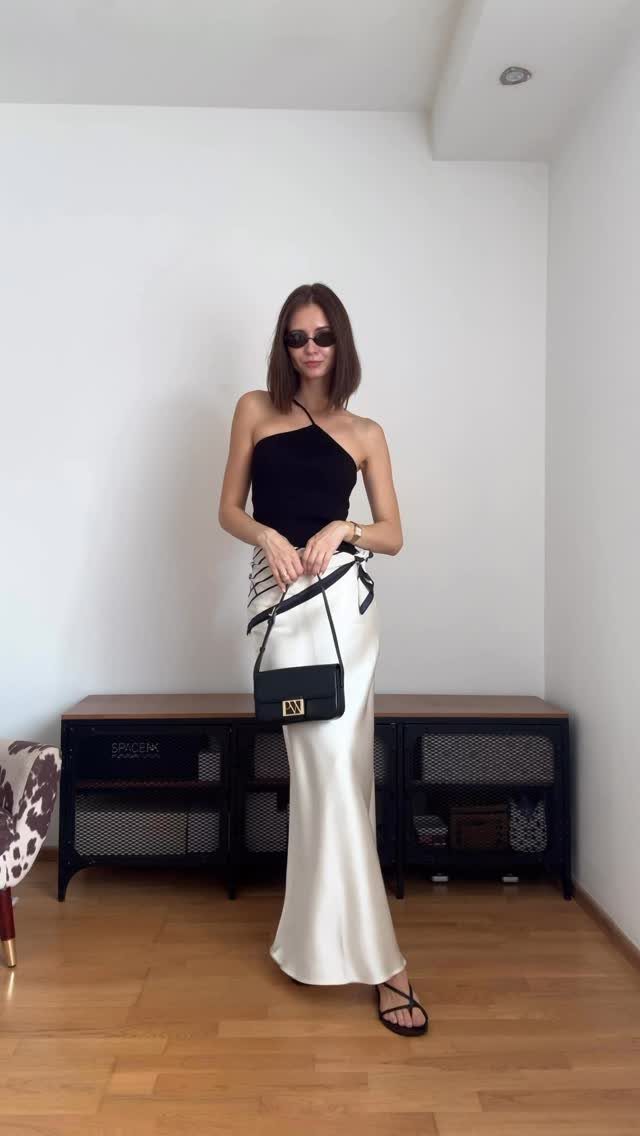 Woman wearing a black halter top, white satin skirt, and black flip-flops standing indoors.