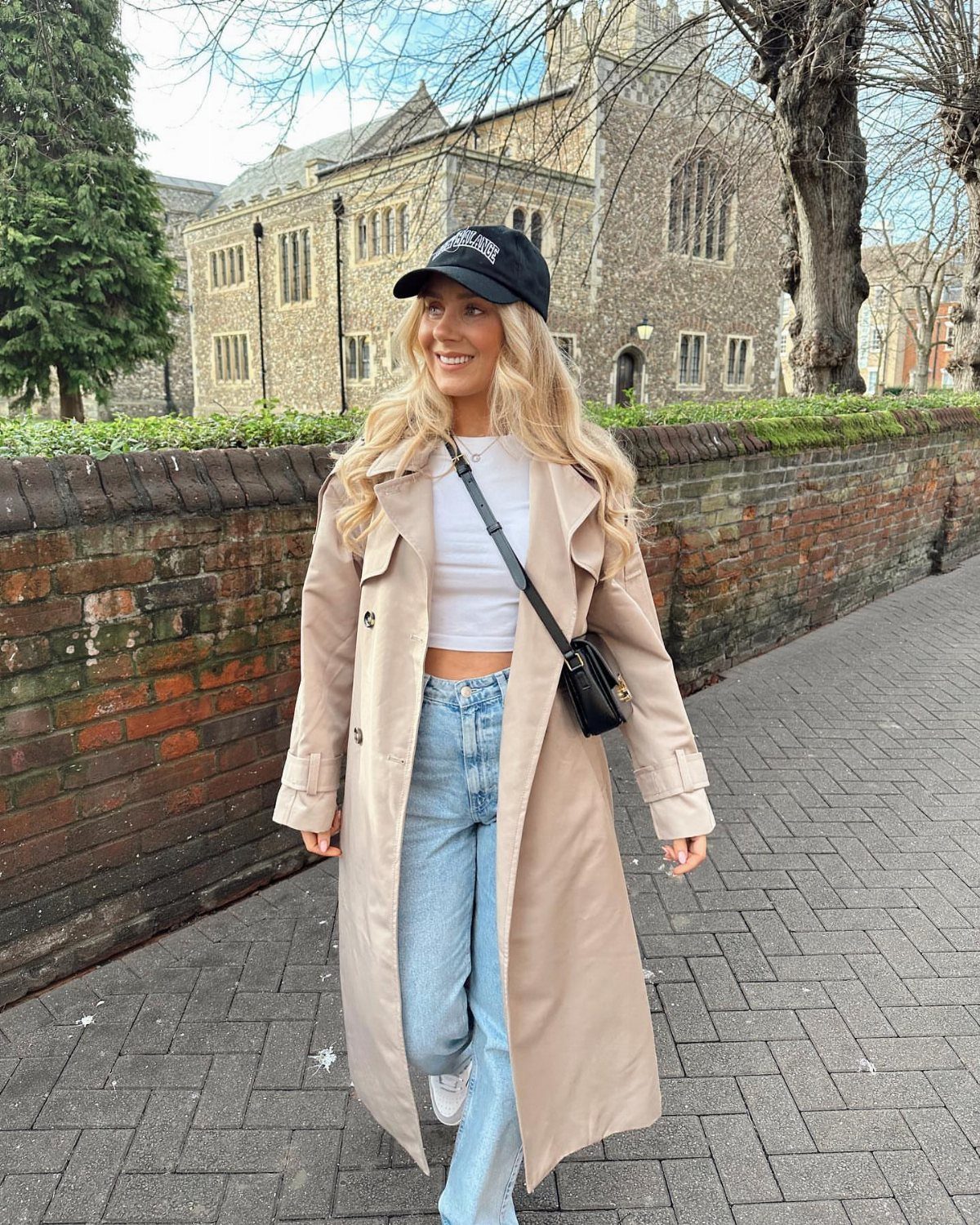 Blonde woman wearing a beige trench coat, white tank top, and blue jeans walking outdoors.