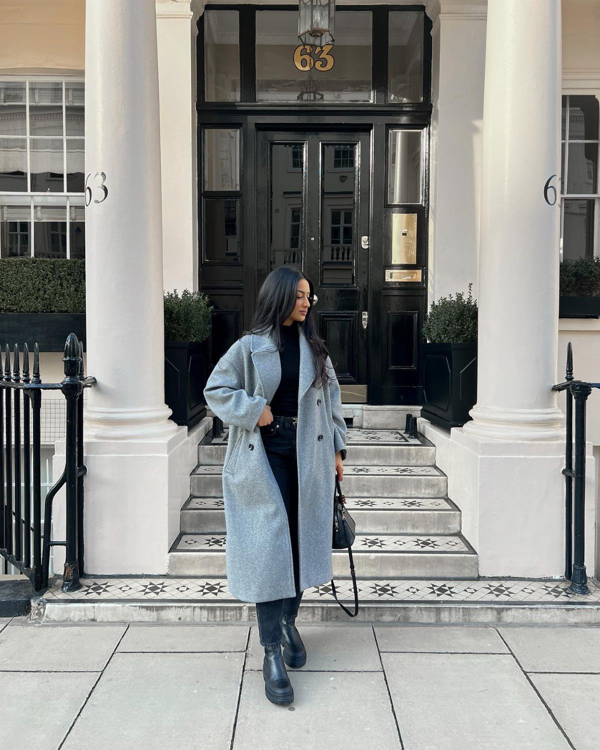 Woman wearing a long grey wool coat and black boots standing before a black door.