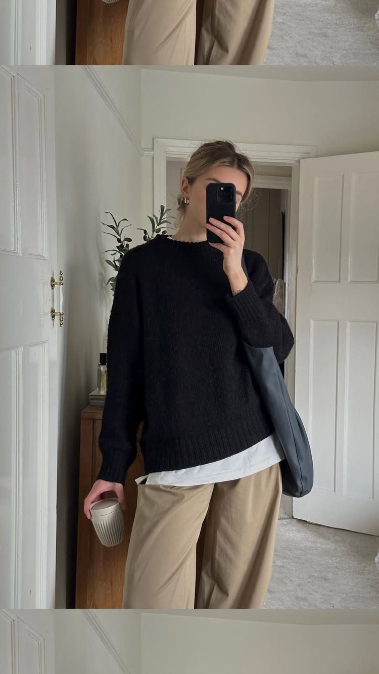 Woman in a black sweater layered over a white shirt and beige trousers taking a selfie.