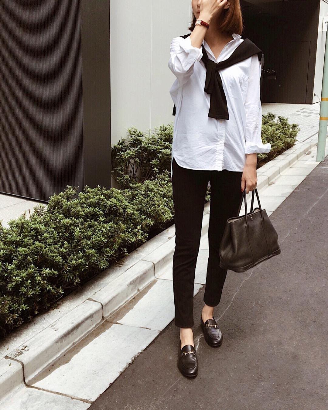 A woman wearing a white shirt, black jeans, and black loafers standing on a sidewalk.