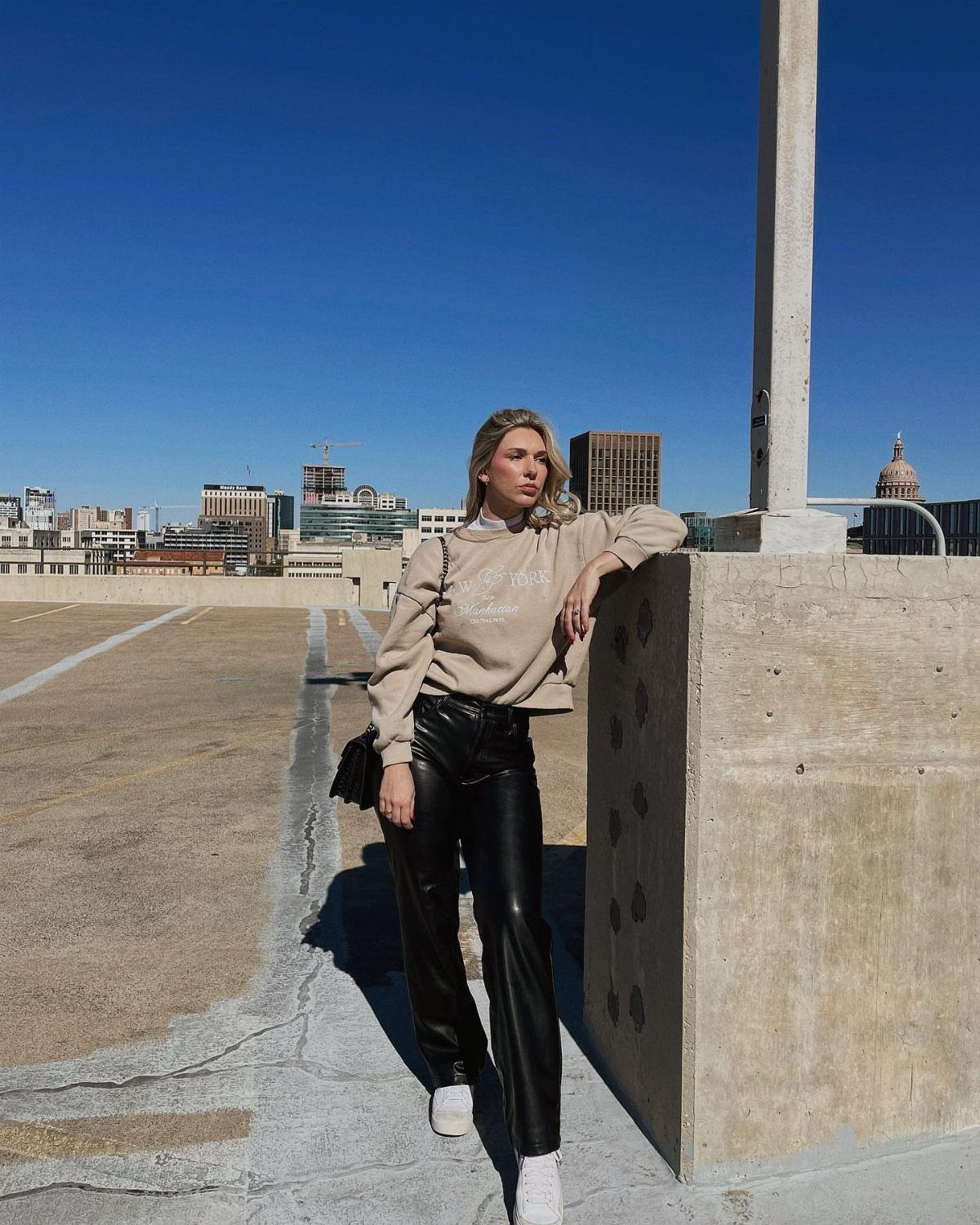 A woman wearing black leather pants and a beige sweatshirt poses on a sunny rooftop.