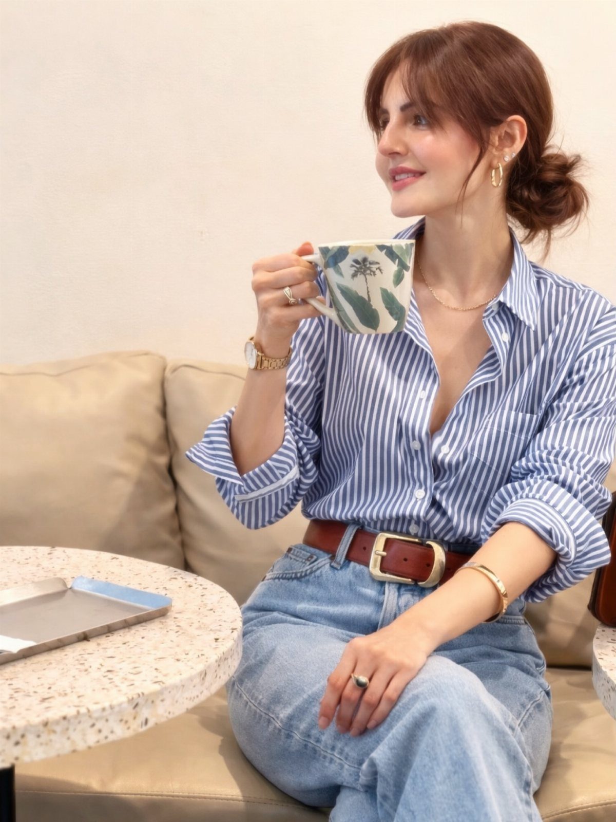 Woman in a striped button-down shirt and blue jeans sitting on a sofa holding a mug.