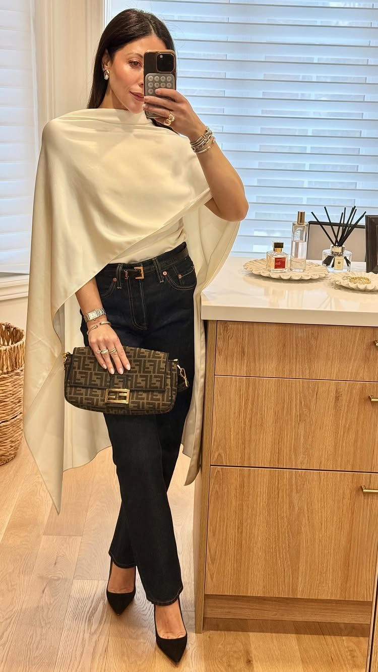 Woman in a cream draped top, dark jeans, and black heels posing for a mirror selfie.