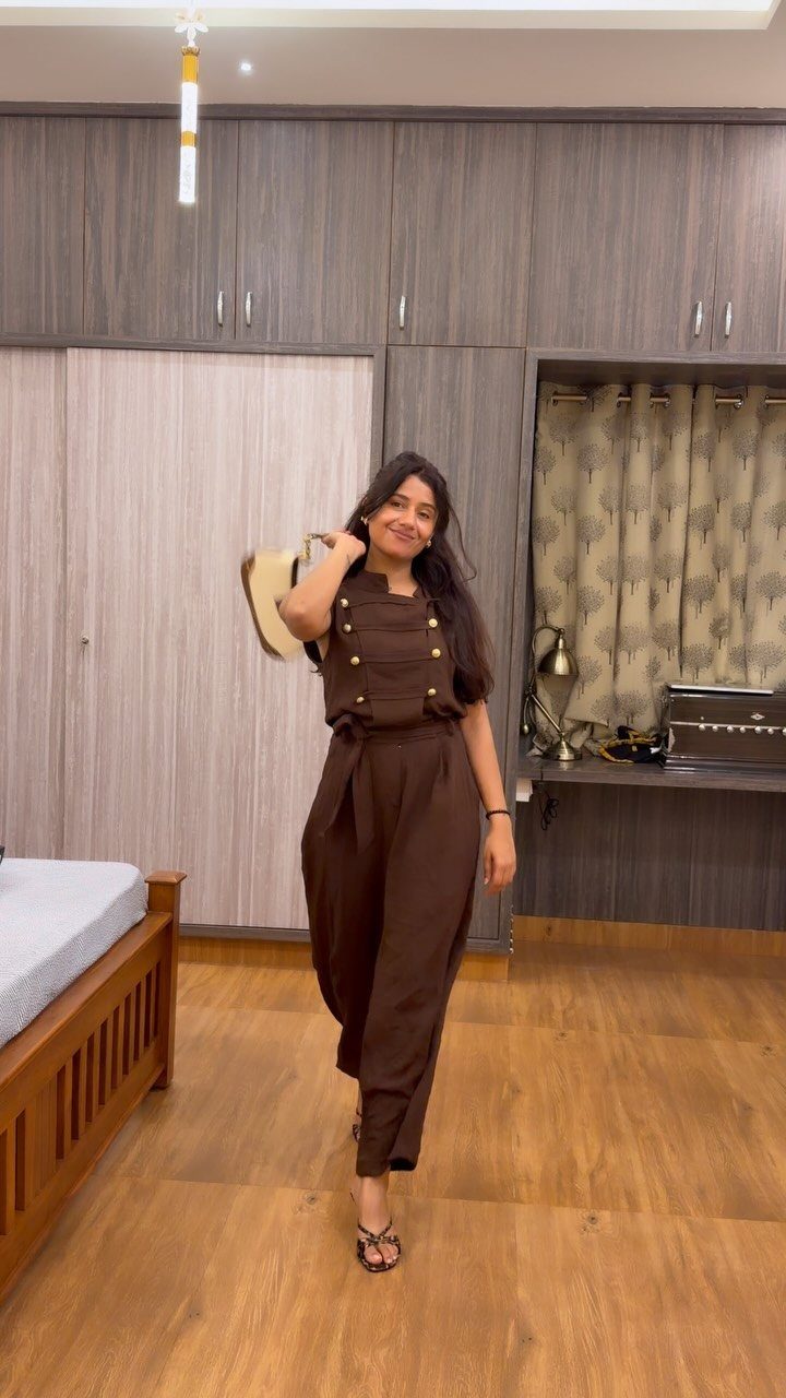 A smiling woman posing in a brown sleeveless jumpsuit and black sandals indoors.
