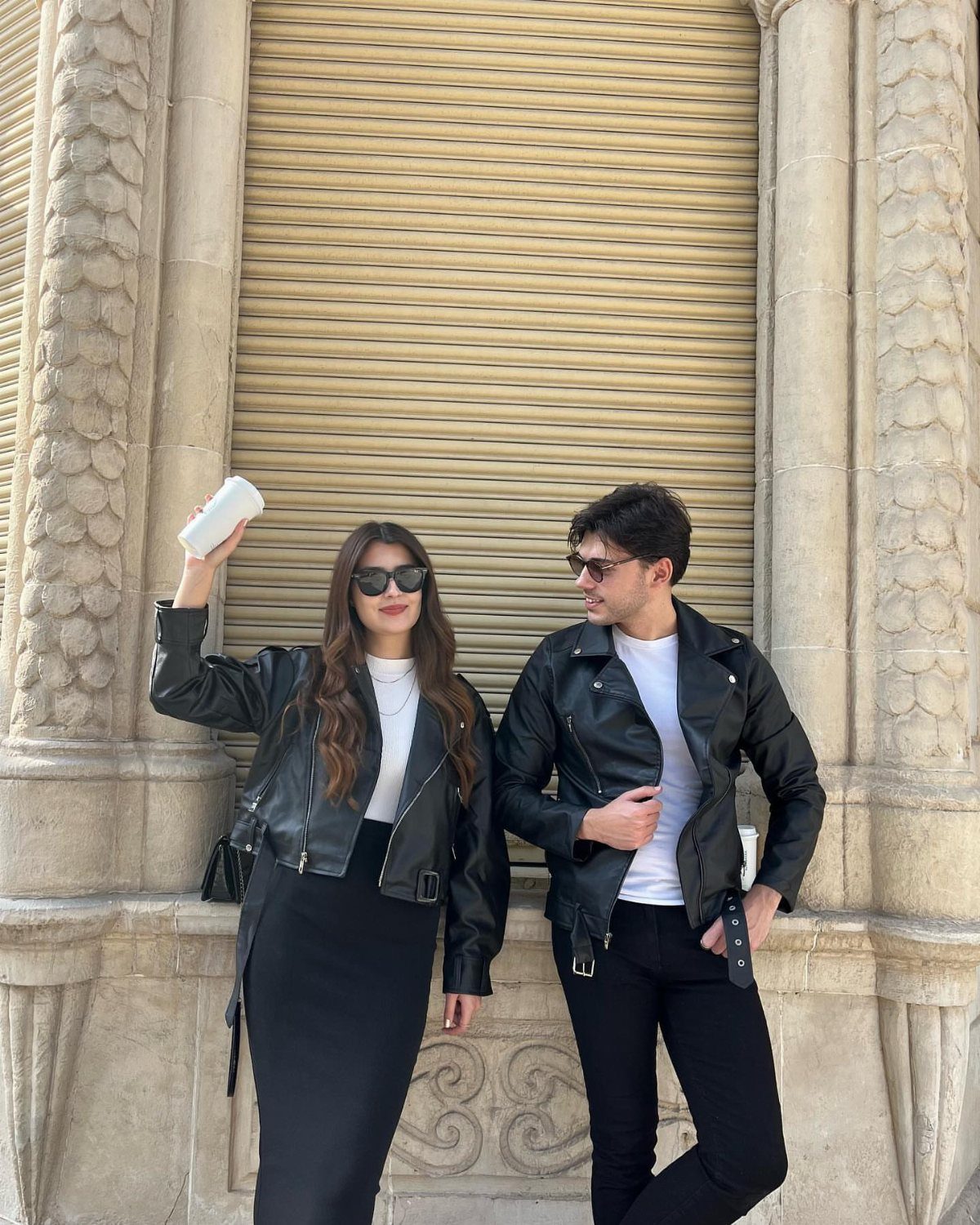 A man and woman posing in black leather jackets and white t-shirts outdoors.