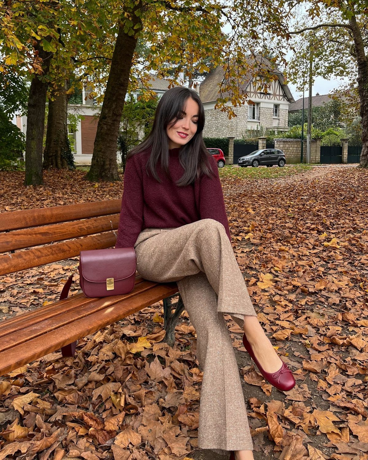 Woman in burgundy sweater and tan trousers sitting on a bench covered in autumn leaves.