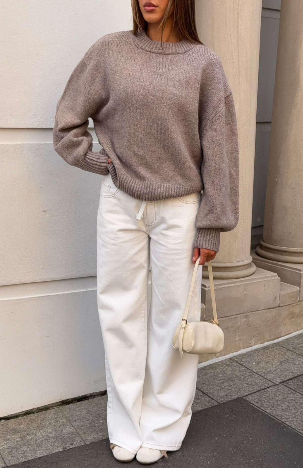 A person wearing a mauve oversized knit sweater and white wide-leg trousers holding a small handbag.