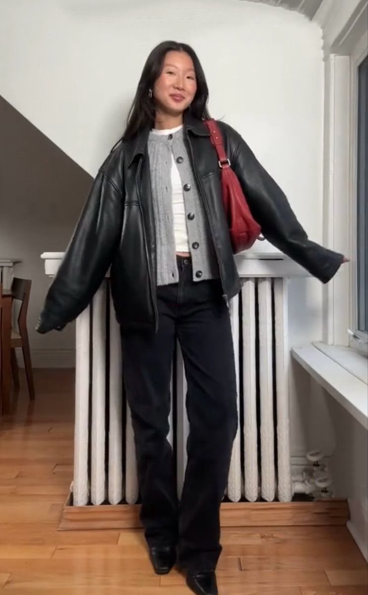 A woman posing in a black leather jacket, grey cardigan, and black trousers.