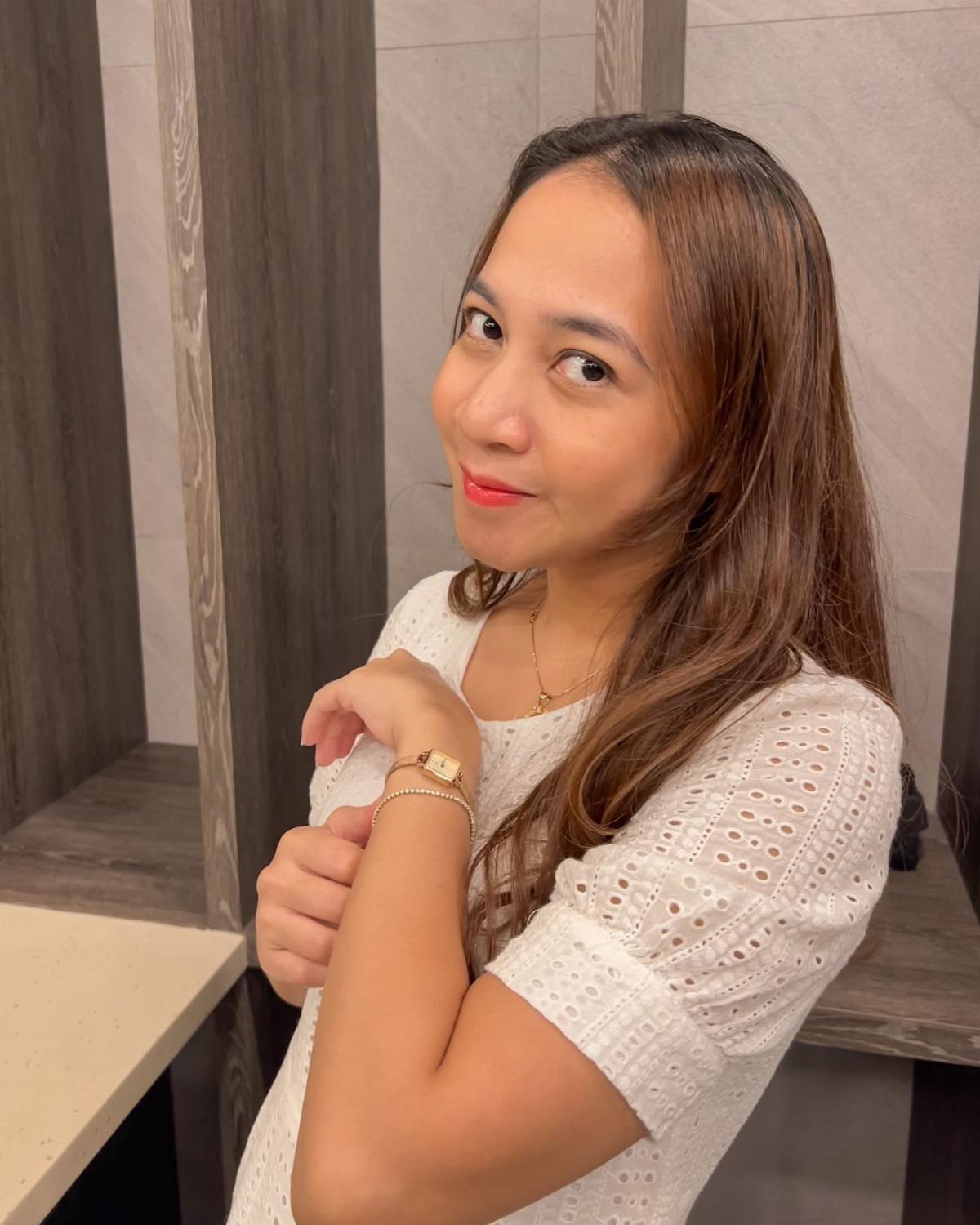 A smiling woman posing while showcasing a gold minimalist watch and bracelet on her wrist.