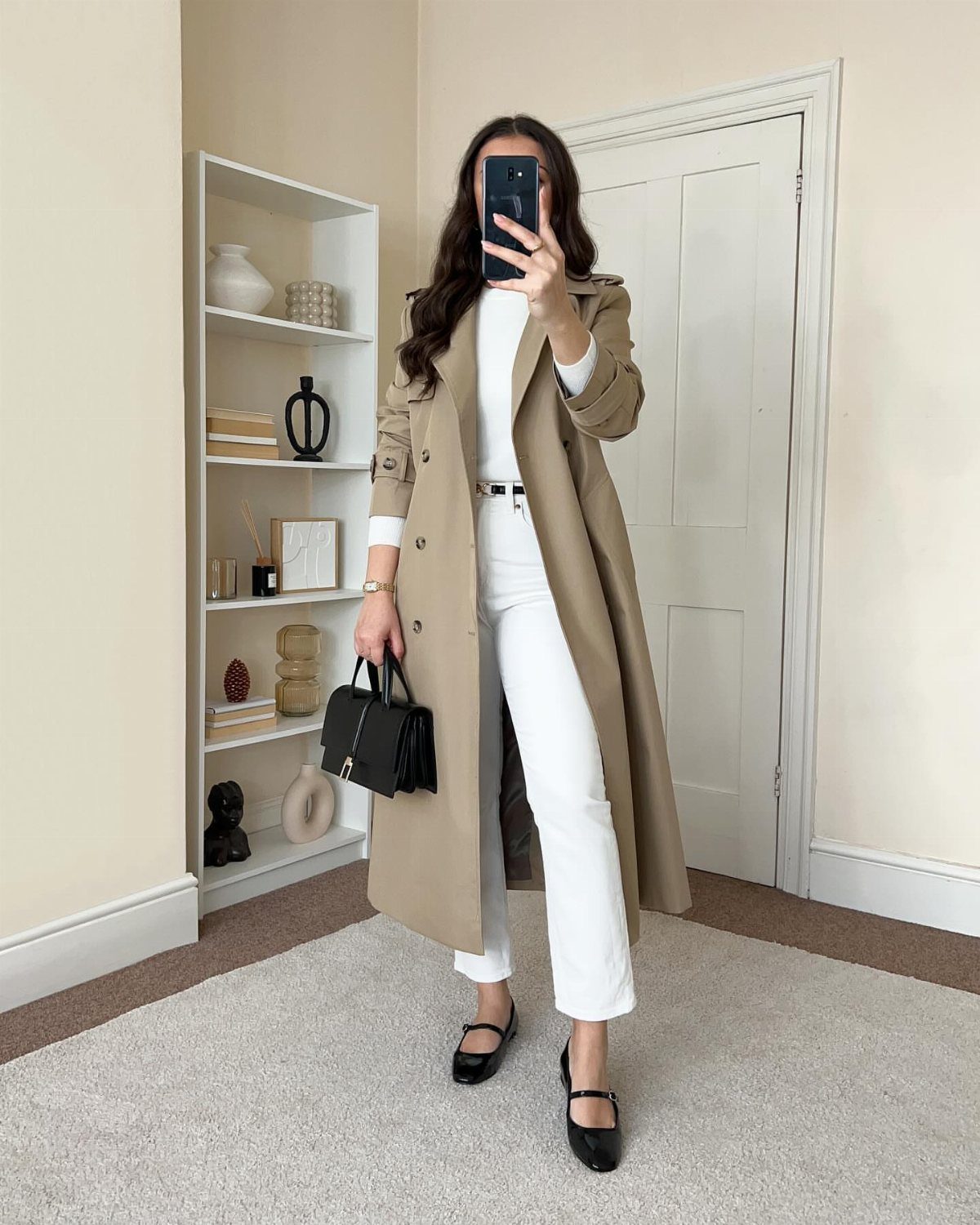 Woman in a long beige trench coat, white trousers, and black flats holding a handbag.
