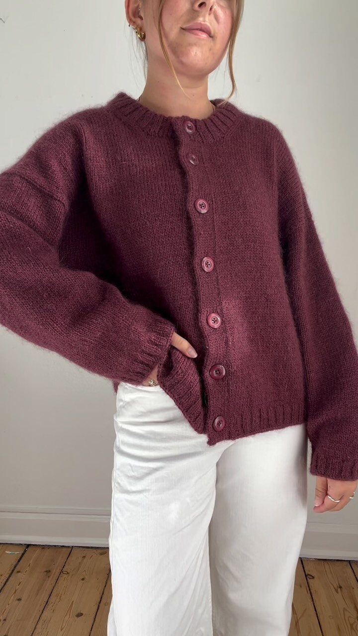 A person wearing an oversized burgundy knit cardigan and white trousers against a white wall.