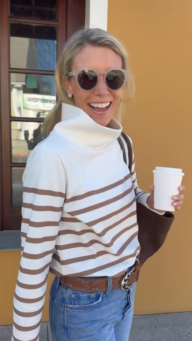 A smiling woman wearing a striped white and brown turtleneck sweater and blue jeans.