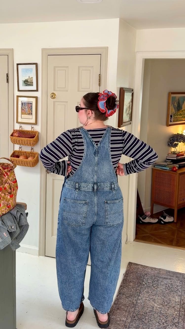 Woman wearing denim overalls, a striped shirt, and brown clogs standing in a room.