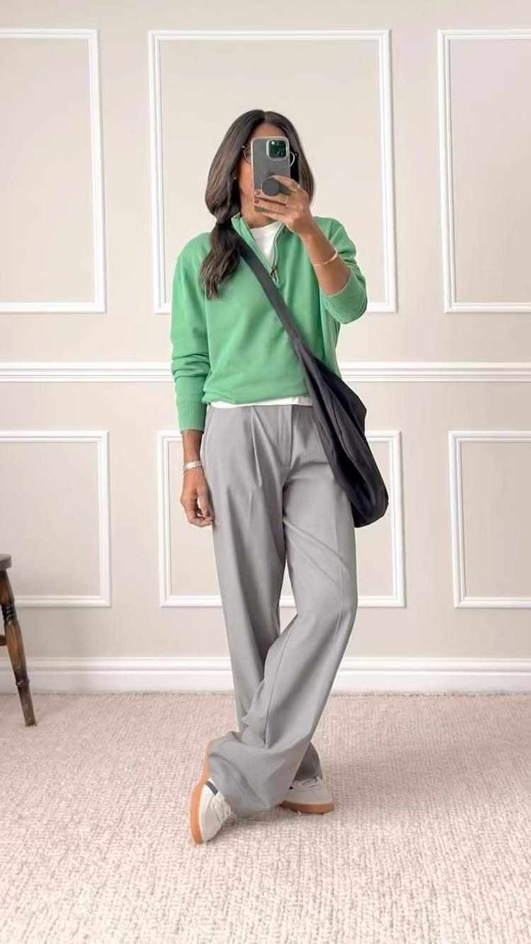 A woman posing in a green pullover and grey wide-leg trousers while taking a mirror selfie.