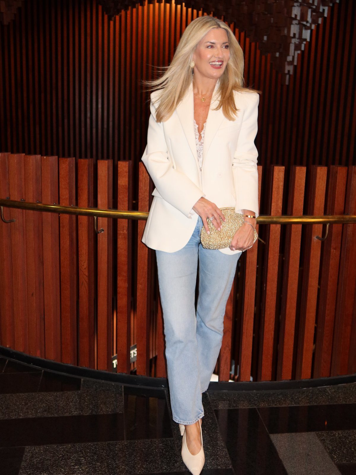 A blonde woman wearing a white blazer and light blue denim jeans standing in front of wooden slats.
