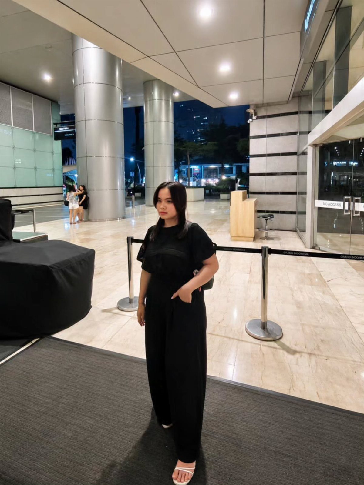 A woman wearing a black short-sleeved jumpsuit and white sandals standing in a modern building lobby.