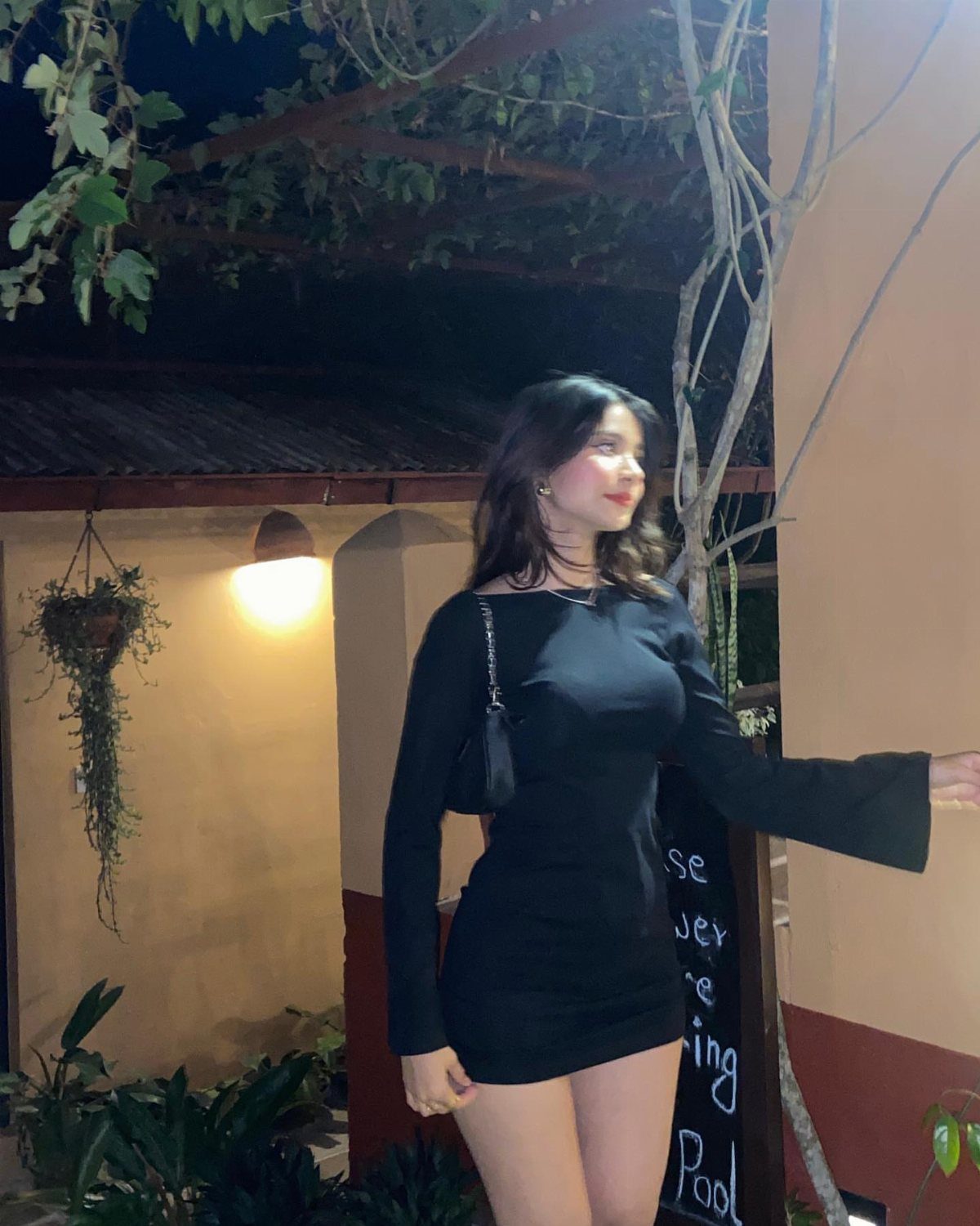A woman in a black long-sleeved minidress standing outdoors at night near greenery.