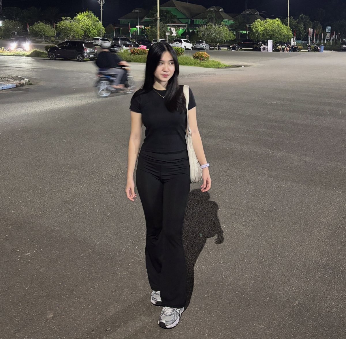 A woman wearing an all-black casual outfit standing on a paved lot at night.