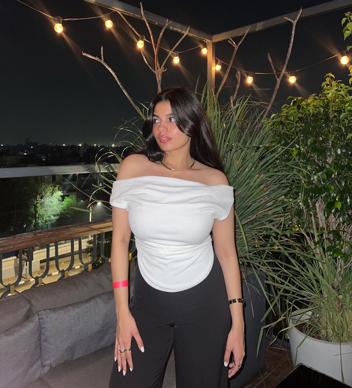 A woman wearing a white off-the-shoulder top and black pants on a rooftop at night.