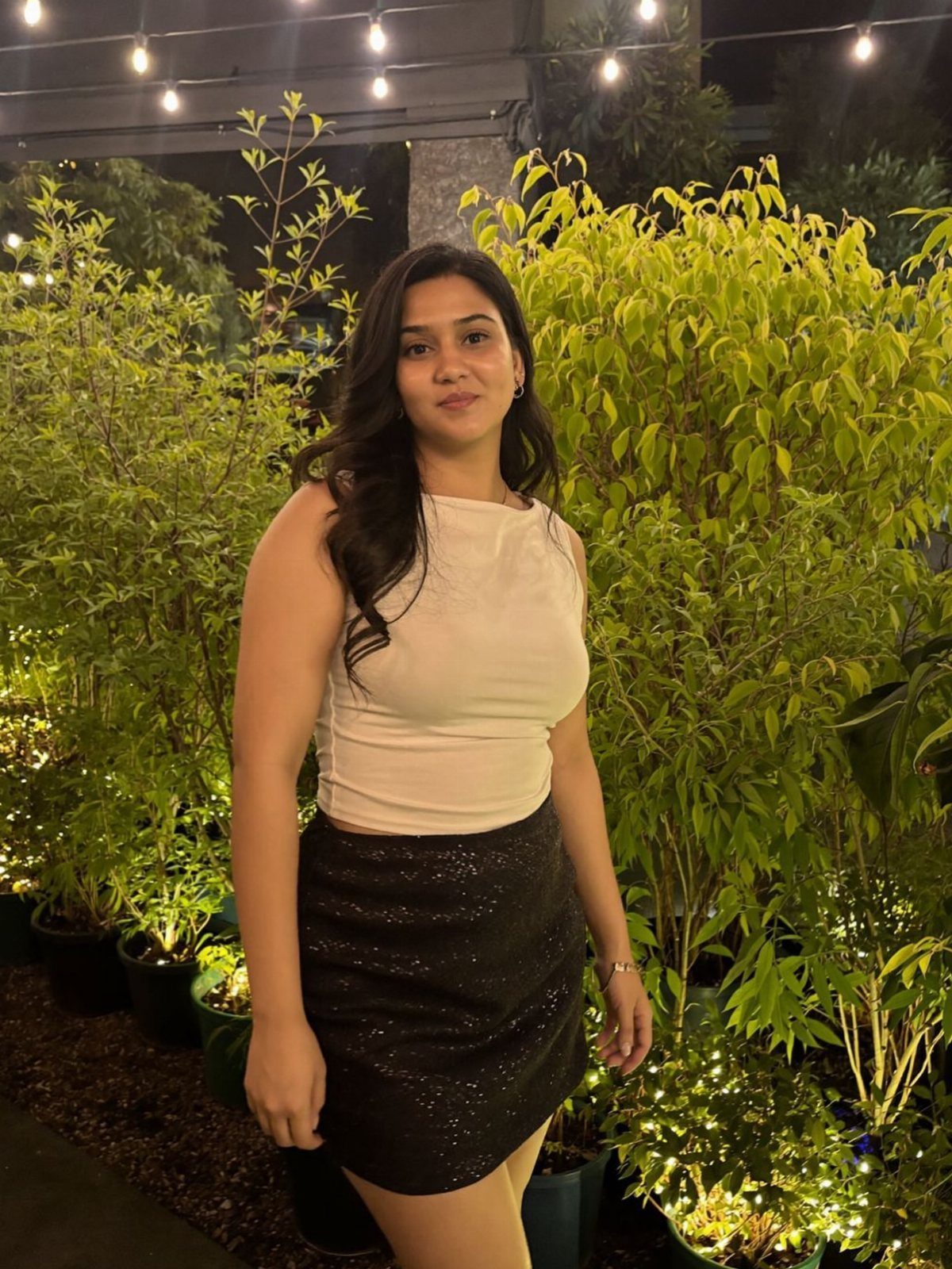 A woman wearing a light crop top and a black sequined skirt standing among green plants.
