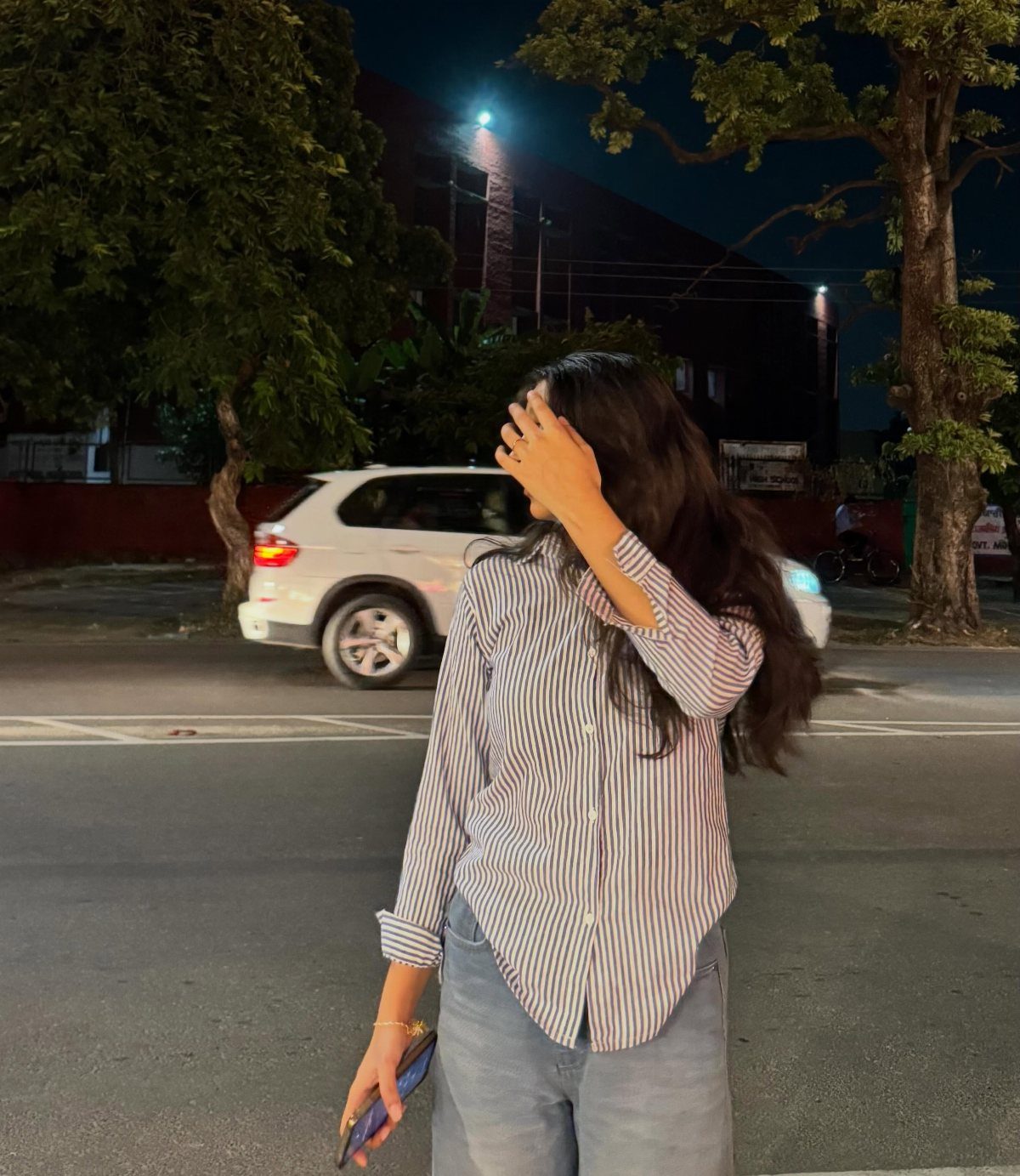 A woman in a striped button-down shirt and blue jeans standing on a street at night.