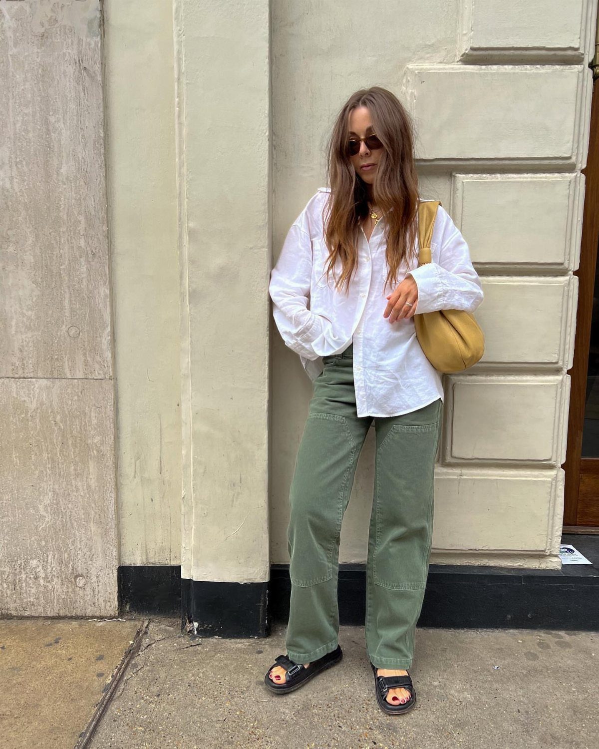 A woman in a white shirt and green trousers posing against a stone wall.