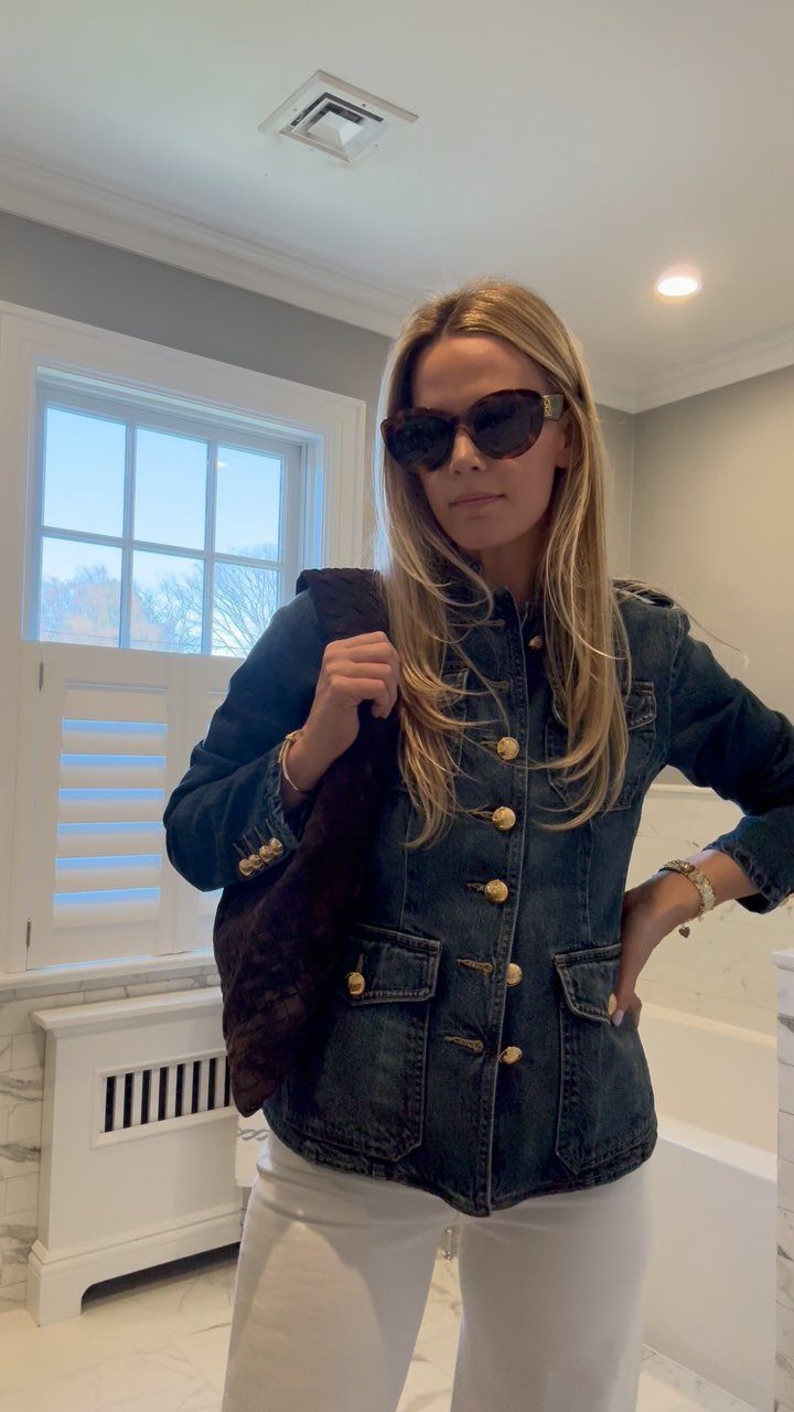 A woman wearing a dark denim jacket with gold buttons and white pants standing indoors.
