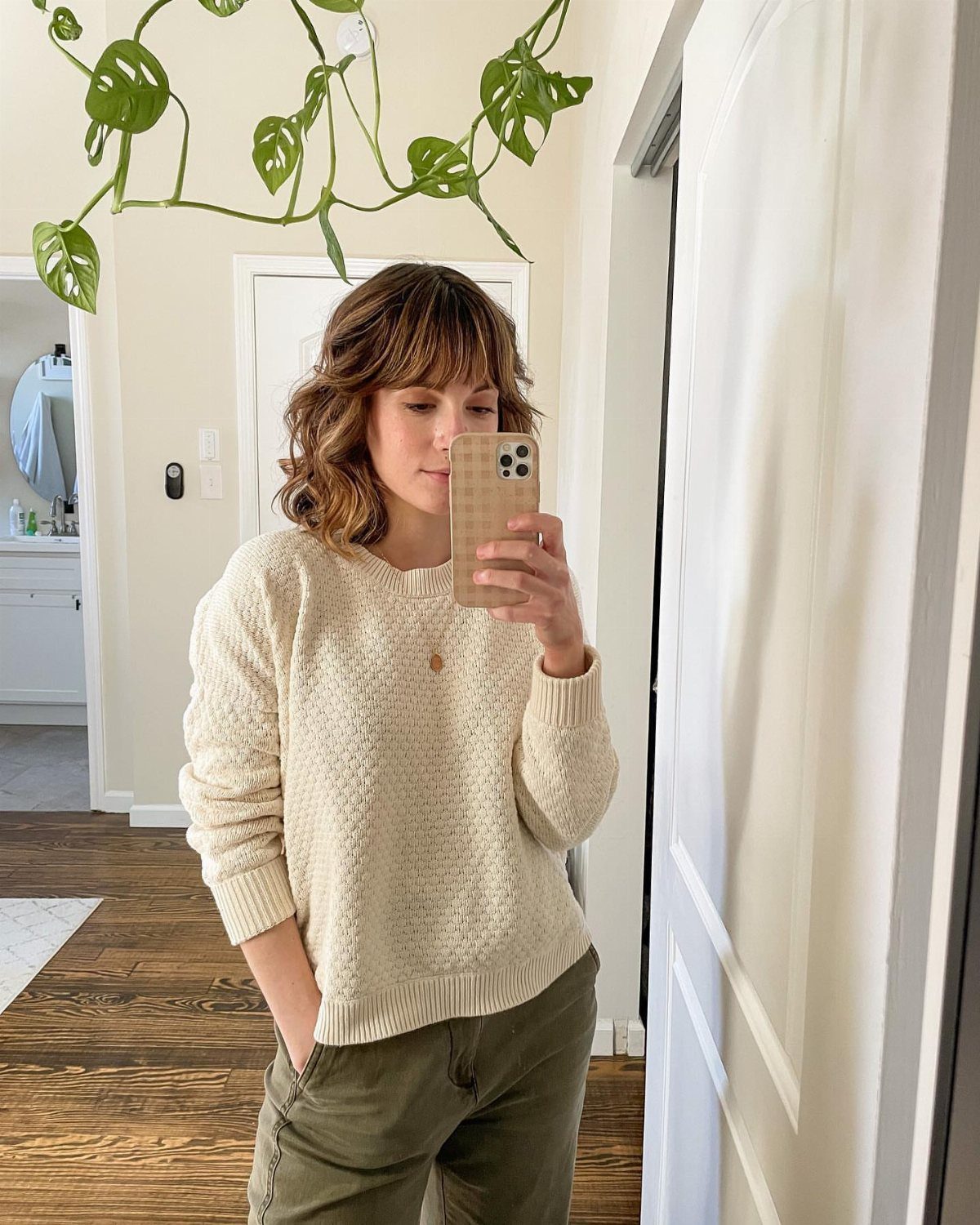 Woman in a cream knit sweater and olive green pants taking a mirror selfie.