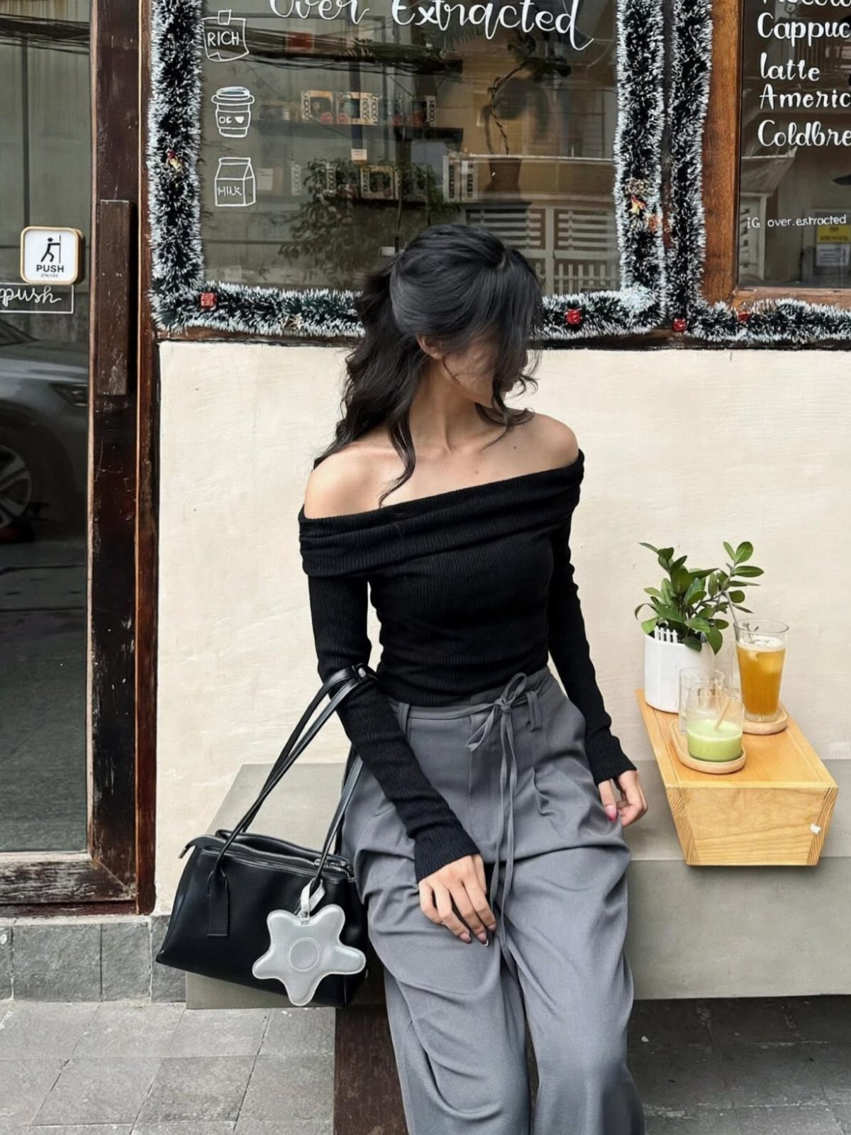 A woman wearing a black off-the-shoulder top and grey trousers sitting on a bench.