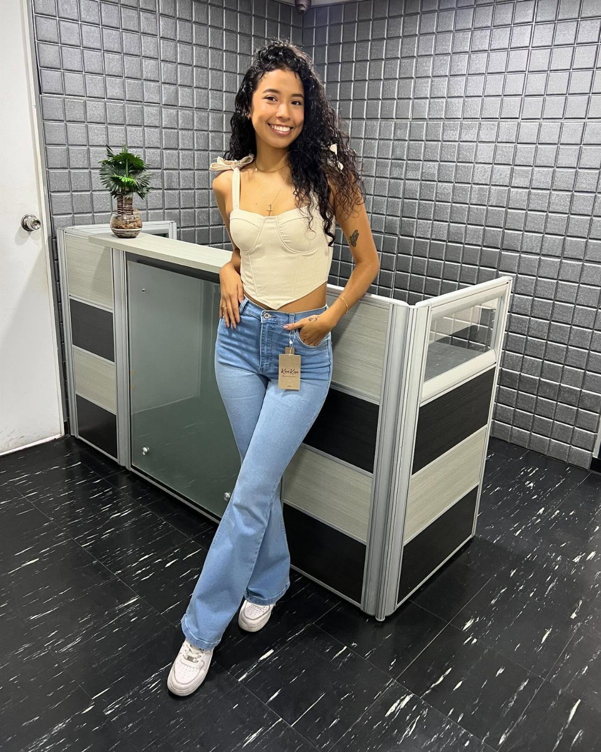 A smiling woman wearing a cream corset top and light blue jeans posing indoors.