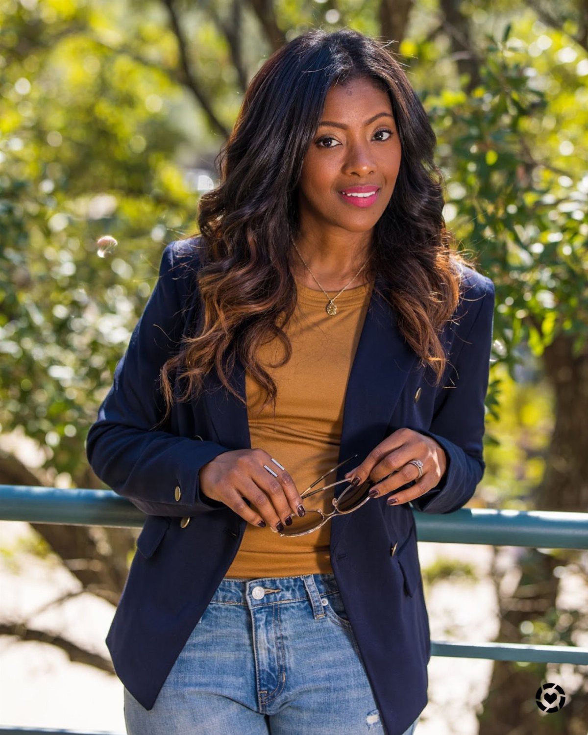 A woman wearing a navy blazer, mustard top, and blue jeans posing outdoors.