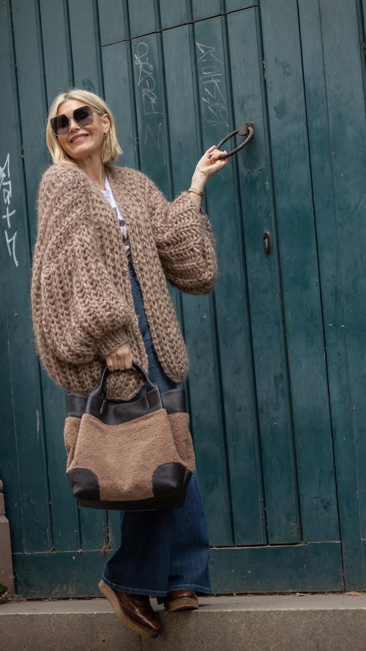 A woman wearing a chunky beige knit cardigan and blue jeans standing by a teal door.