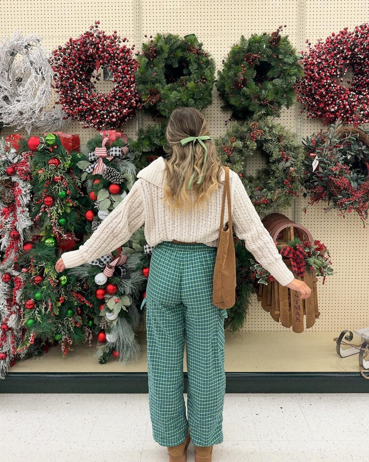 Woman in a cream sweater and green plaid trousers standing before festive Christmas wreaths.