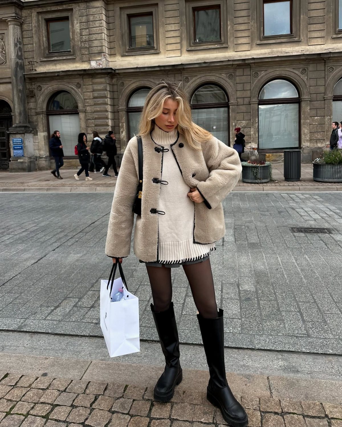 Woman in a cream sweater dress, fuzzy teddy jacket, black tights, and tall black boots.