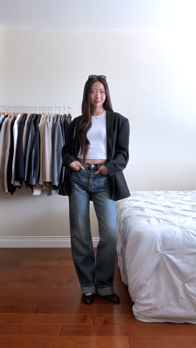 A woman wearing a black blazer, white top, blue jeans, and black ballet flats.