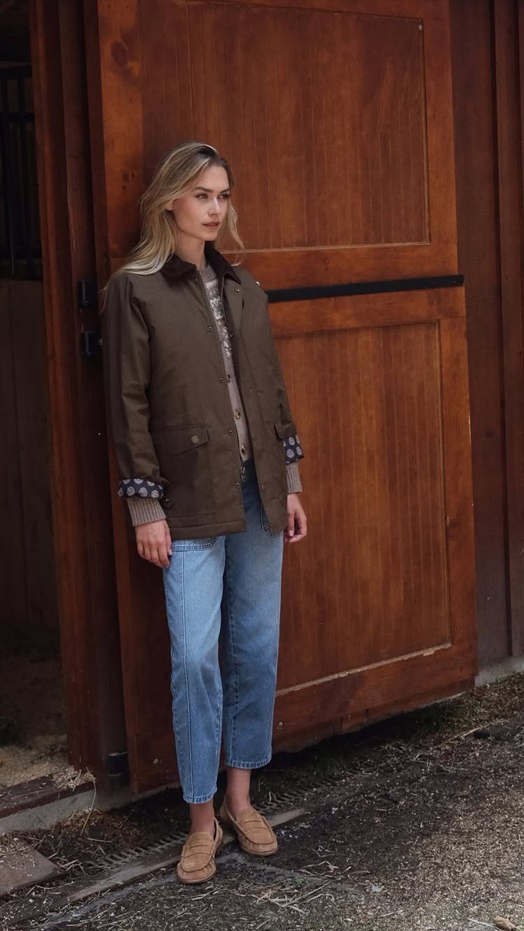Woman wearing a brown barn jacket, gray sweater, and blue jeans leaning against a wooden door.