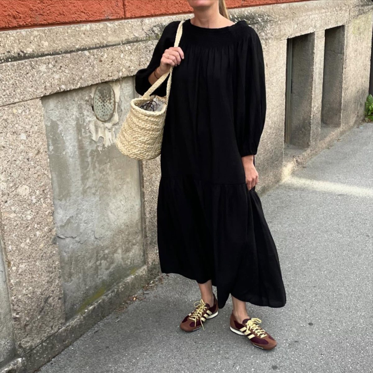 A person wearing a black tiered maxi dress and burgundy sneakers while carrying a straw bag.