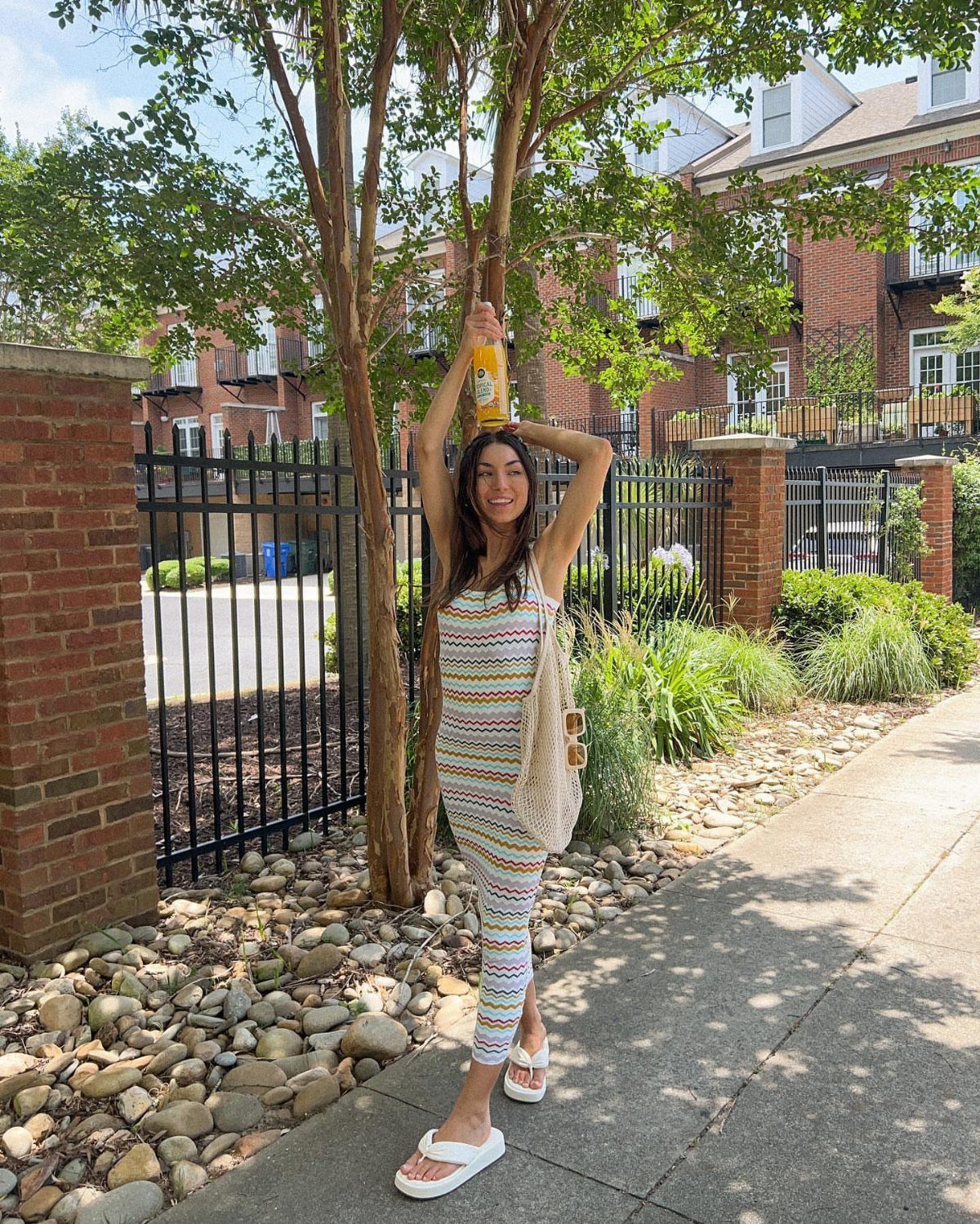 A smiling woman in a long striped knit dress and white sandals standing by a tree.