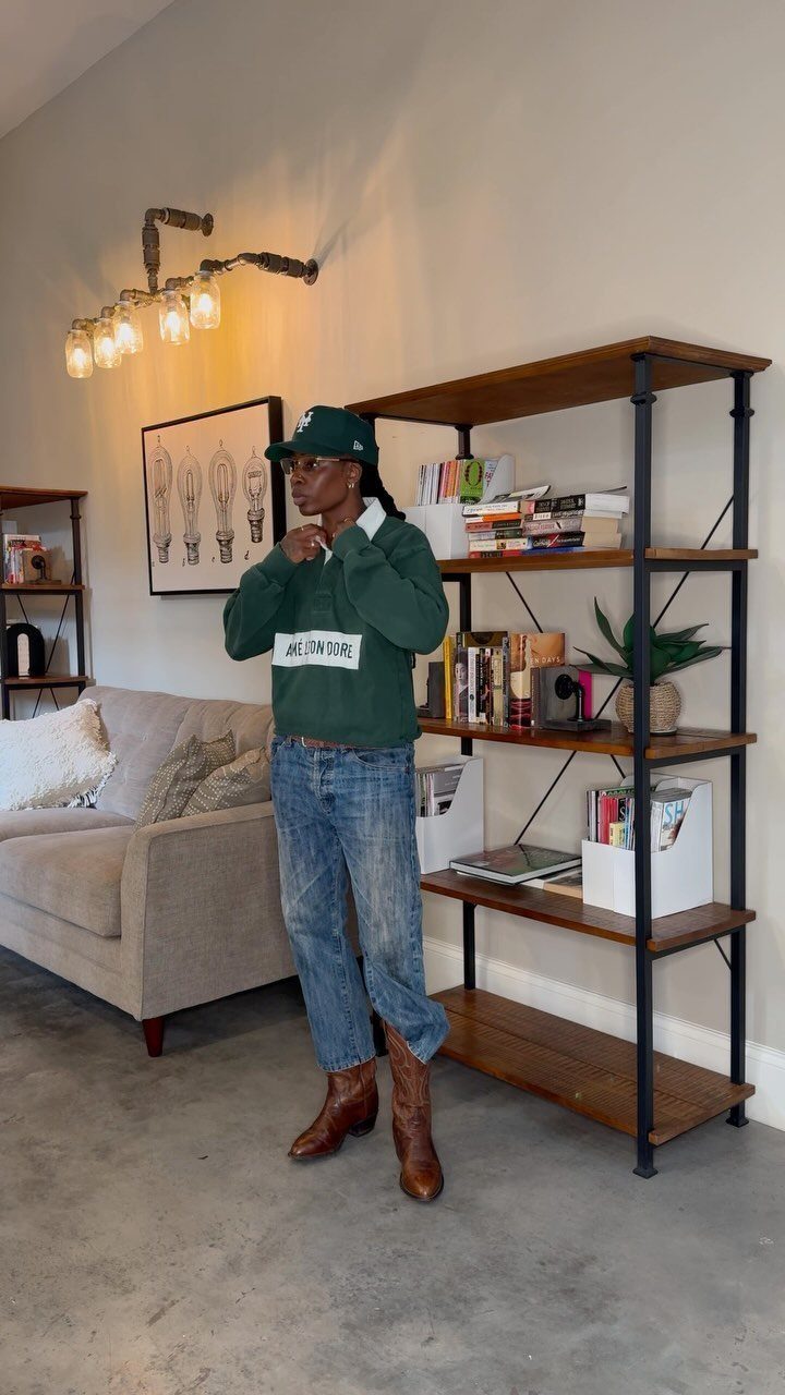 A person wearing a green sweatshirt, blue jeans, brown boots, and a green baseball cap.