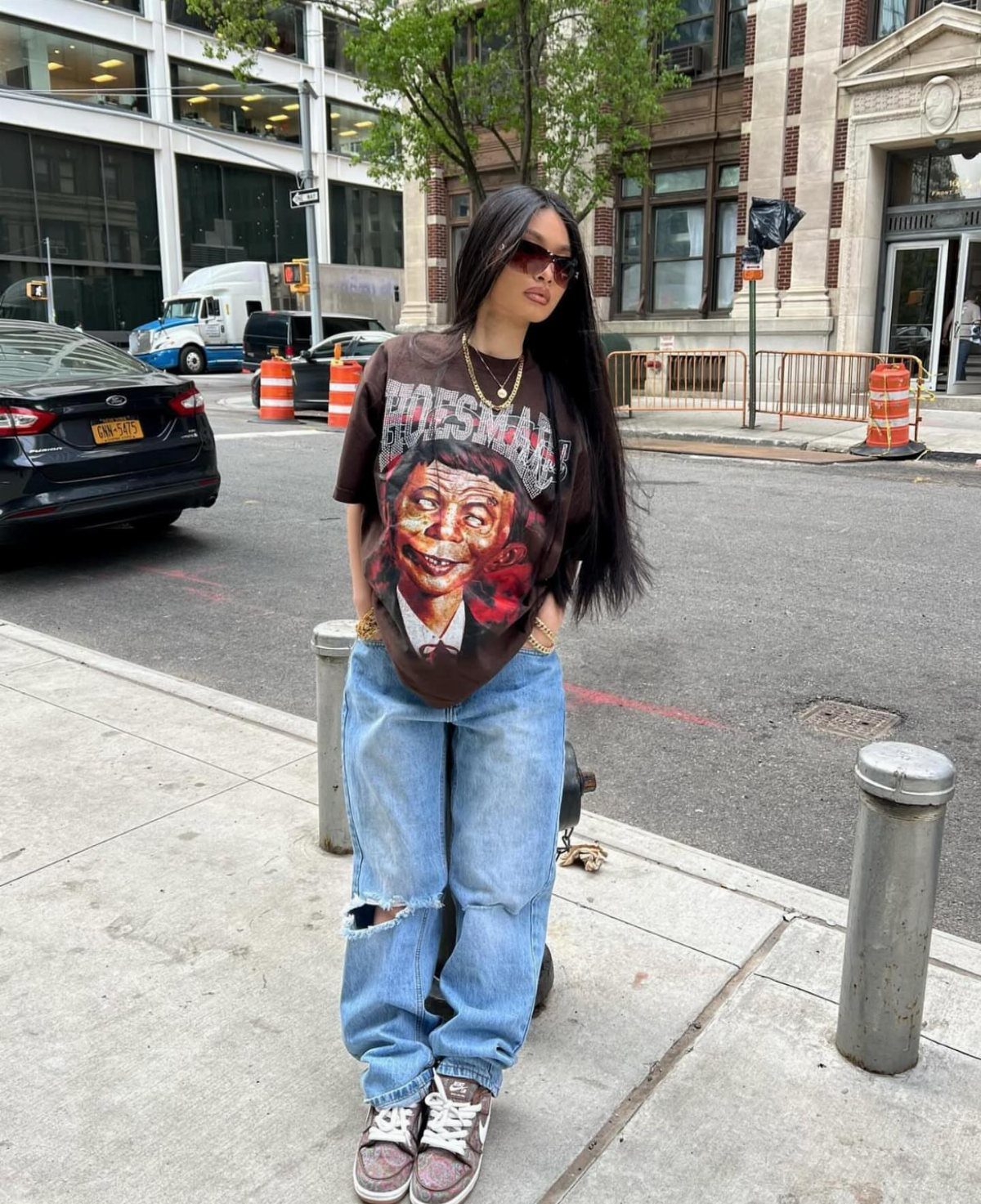 Woman wearing a brown graphic t-shirt, ripped blue jeans, and sneakers standing on a city sidewalk.