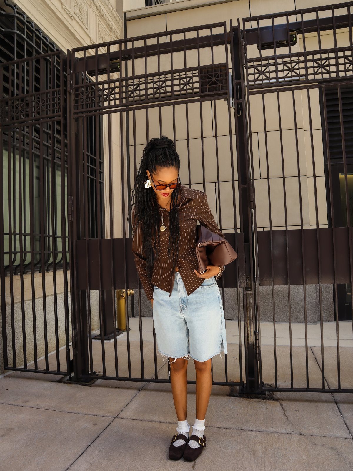 Woman in denim shorts and black loafers standing before a large black metal gate.