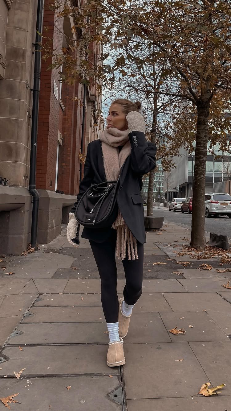 Woman walking on a city sidewalk wearing a black blazer, beige scarf, and shearling boots.