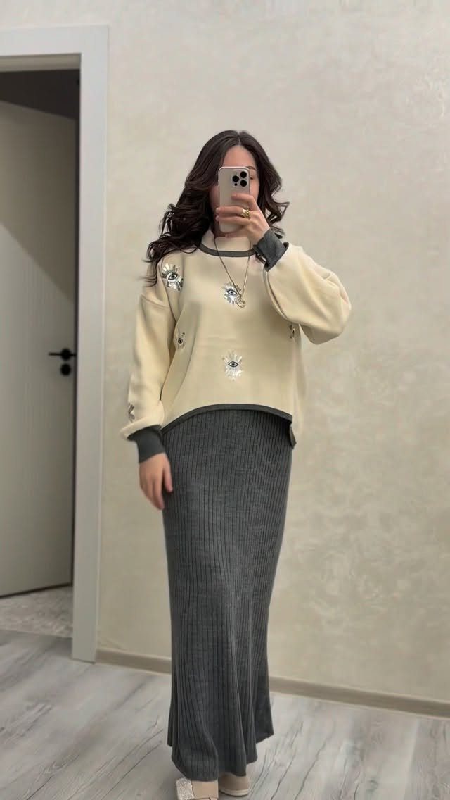 Woman in a cream sweater and long grey ribbed skirt taking a mirror selfie.