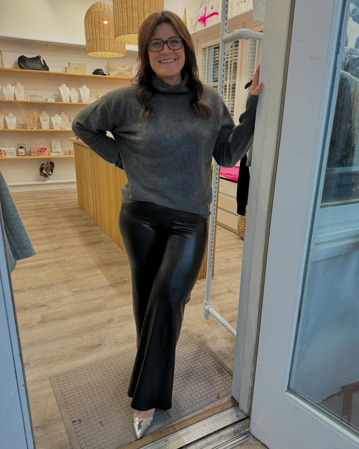 A woman posing in a grey turtleneck sweater and black faux leather pants inside a boutique.