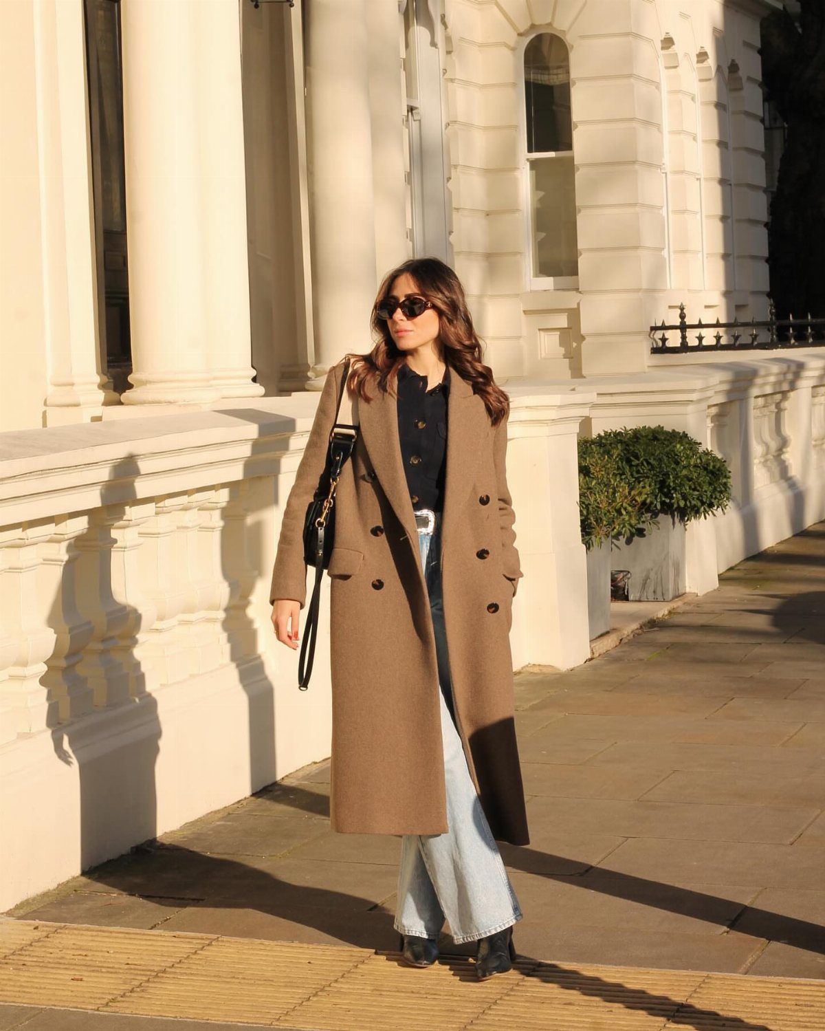Woman wearing a long tan wool coat and wide-leg jeans standing on a sunny sidewalk.