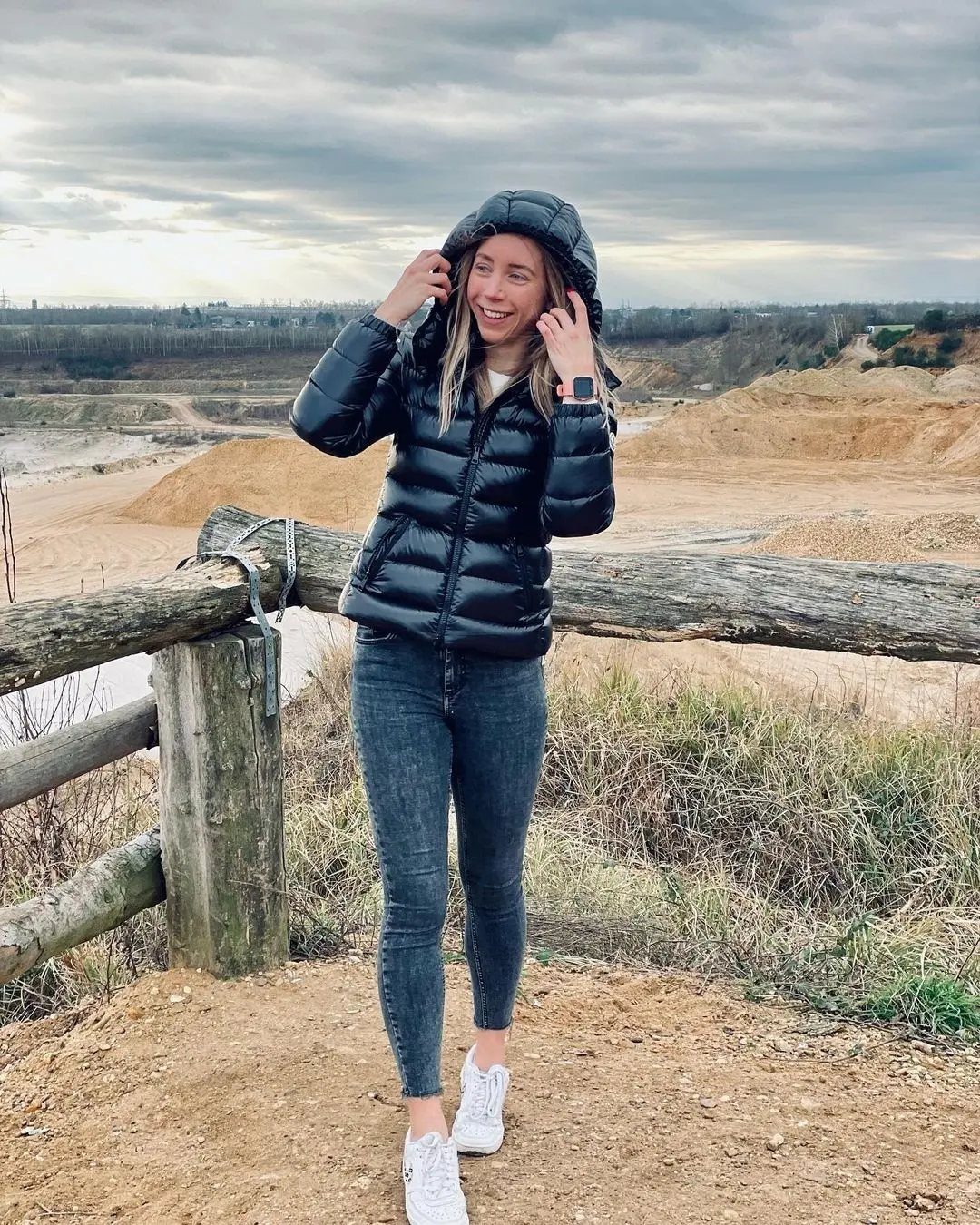 A smiling woman wearing a black puffer jacket and blue jeans standing outdoors.