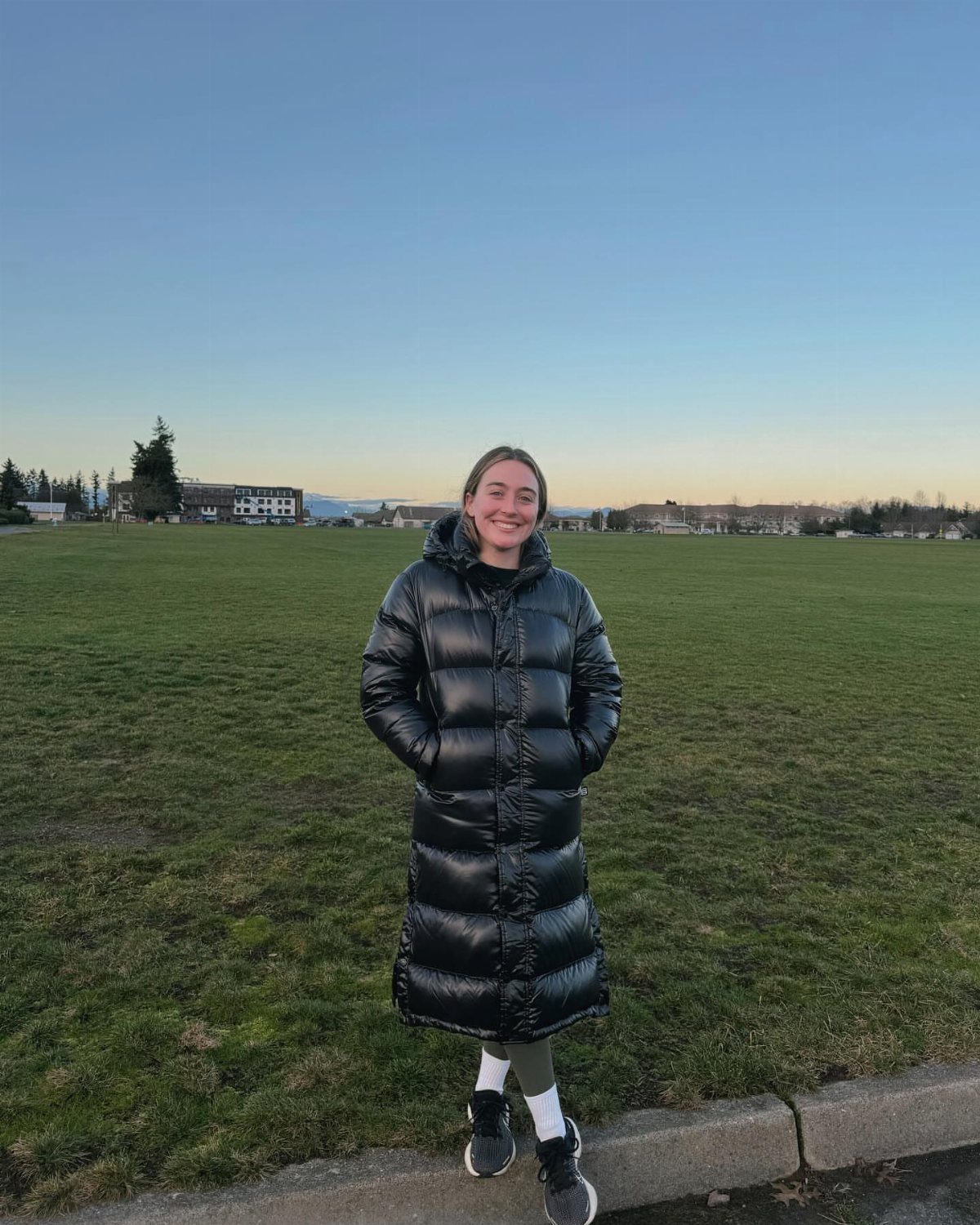 A smiling woman wearing a long black puffer jacket and grey leggings in a grassy field.